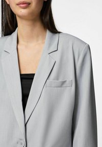 Gaiši pelēks žakets ar gludu tekstūru, ar vienu pogu, ar iedobtiem mēteļa galiem un krūšu kabatu. Uzvilkts virs melna topa.