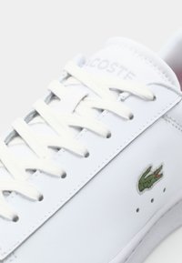 Valge nahast tenniseking, millel on lapikud valged paelad, Lacoste logo, roheline tikitud krokodill oraz külgfreesitud detailide juurde.