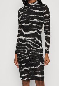 Robe noire à manches longues cintrée avec des rayures abstraites en zèbre blanches et un tissu texturé, portée par une personne debout avec des cheveux tressés.