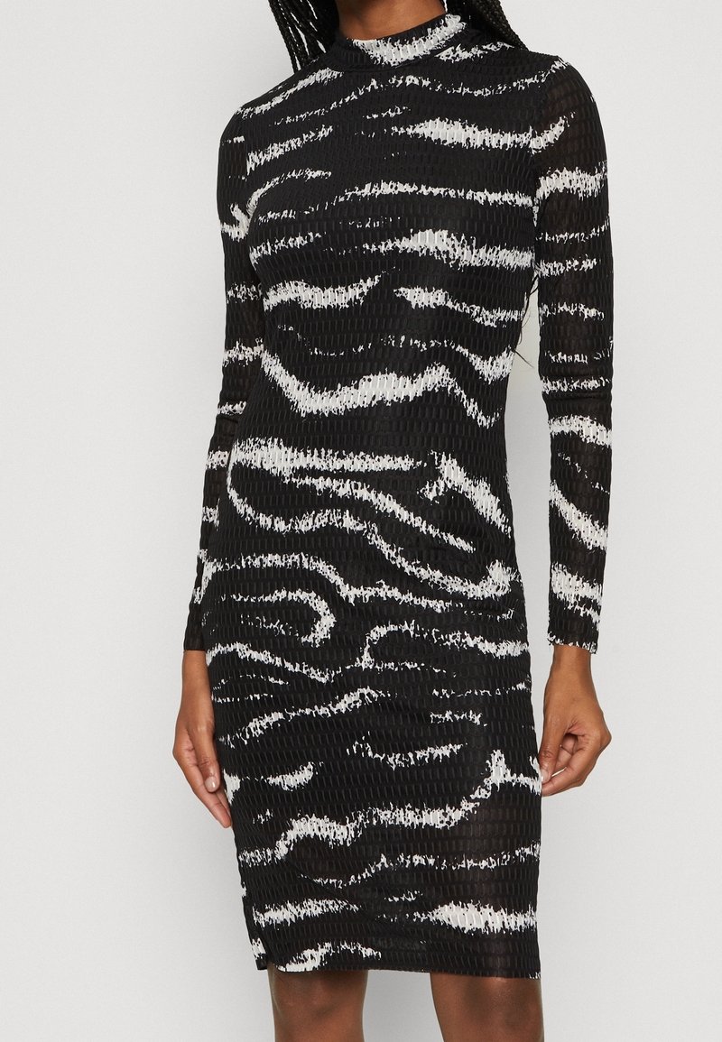 Robe noire à manches longues cintrée avec des rayures abstraites en zèbre blanches et un tissu texturé, portée par une personne debout avec des cheveux tressés.