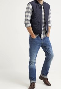 Navy quilted vest over a gray and white checkered shirt, paired with blue jeans and brown leather boots, standing against a light background.