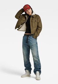 Olijfgroene cropped jacket met schapenvachtvoering, zwarte top, verouderde blauwe spijkerbroek en witte high-top sneakers. Rode pet met logo.