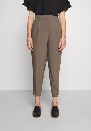 Person trägt hochtaillierte, zulaufende Hose in Taupe, schwarzes kurzärmeliges Rüschenoberteil und schwarze Slip-On-Schuhe und steht vor einem einfarbigen Hintergrund.