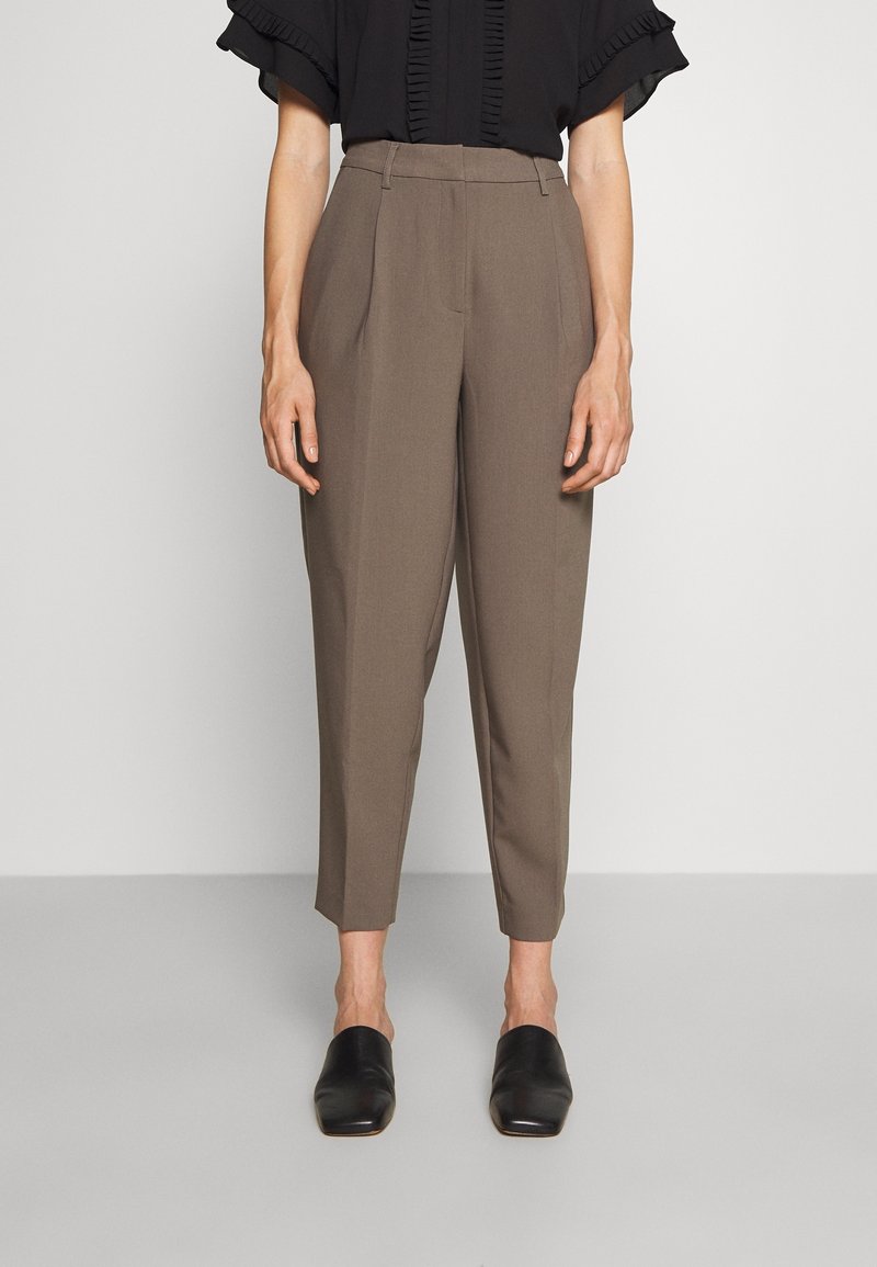 Personne portant un pantalon taupe taille haute et fuselé, un top noir à manches courtes volantées et des chaussures noires sans lacets, debout devant un fond uni.