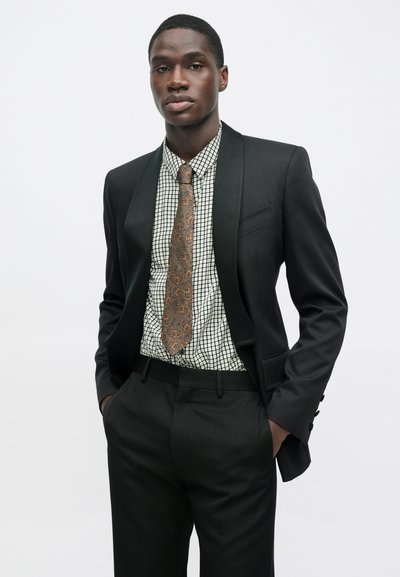 Čierny smoking so zeleným saténovým klopom, doplnený košeľou s mriežkovým vzorom a hnedou paisley kravatou. Strih na mieru s bočnými vreckami.