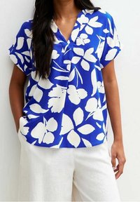 Blouse à manches courtes et col en V en tissu bleu vif avec un motif floral blanc. Portée avec un pantalon ample de couleur claire.