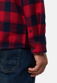 Chemise en flanelle à carreaux rouge et marine avec des poignets boutonnés à deux boutons, portée avec un jean en denim foncé.