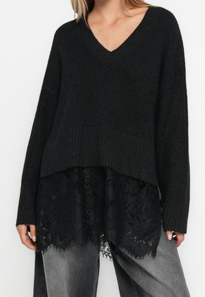 Femme portant un pull en maille noir à col en V superposé à un top en dentelle noire, associé à un jean gris ample délavé.