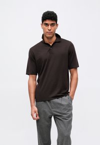 Temno rjav polo majica s kratkimi rokavi in ovratnikom, izdelana iz teksturiranega materiala, v kombinaciji s svetlo sivimi krojenimi hlačami.