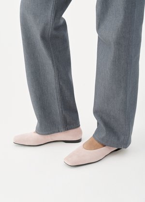 Άτομο που φοράει γκρί τζιν παντελόνι και ανοιχτό ροζ σουέντ επίπεδα παπούτσια, στέκεται πάνω σε λευκή επιφάνεια.