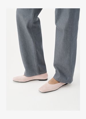 Άτομο που φοράει γκρί τζιν παντελόνι και ανοιχτό ροζ σουέντ επίπεδα παπούτσια, στέκεται πάνω σε λευκή επιφάνεια.