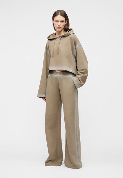 Mujer de pie que lleva una sudadera con capucha beige de gran tamaño y pantalones de pierna ancha con logotipos "OFF", una mano en el bolsillo, sobre un fondo claro y liso.