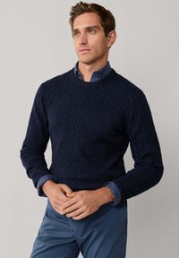 Marineblauwe kabelgebreide trui met ronde nek, textuurpatroon en geribde manchetten, gedragen over een blauw geruit shirt. Gecombineerd met blauwe broek.