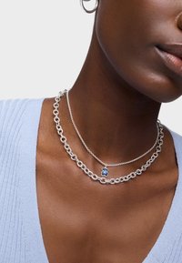 Srebrny naszyjnik łańcuszkowy nałożony na inny, jaśniejszy łańcuszek, z niebieskim zawieszką w kształcie misia. Widoczna teksturowana skóra oraz jasnoniebieska odzież.