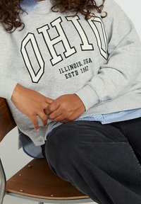 Grå sweatshirt med "OHIO" i fet svart text, lagd över en blå och vit randig skjorta, tillsammans med mörka byxor. Sitter på en trästol.