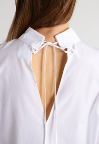 White shirt with open back tied at the neck, showing a woman's upper back and brown hair falling over the collar.