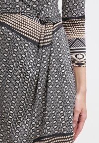 Gros plan sur une robe noire et beige à motifs géométriques et floraux, avec une taille nouée et une main reposant à côté de la hanche.