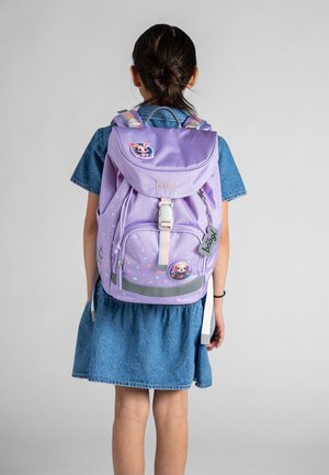 Mädchen in einem Jeanskleid mit einem lavendelfarbenen Rucksack, verziert mit Tier-Patches, steht vor einem einfarbigen grauen Hintergrund.