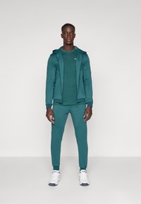 Teal idrettsdrakt bestående av en glidelåsjakke med lommer og matchende tapered bukser, kombinert med hvite treningssko.