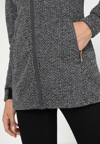 Strikket langærmet jakke i grå og sort med lynlås, sidelommer og et struktureret mønster. Har sort kant omkring ærmerne.