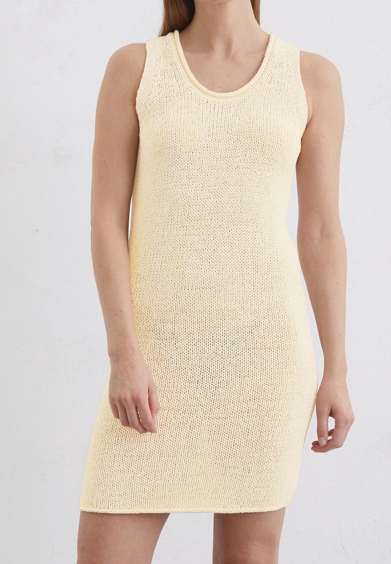 Robe sans manches, longueur au genou, tricotée de couleur jaune pâle, portée par une personne debout devant un fond clair uni.