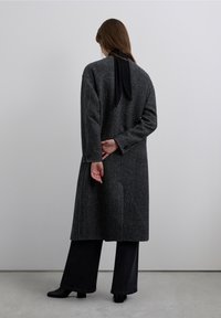 Cappotto lungo a spina di pesce nero e grigio con una silhouette ampia, cuciture dettagliate, grandi bottoni, abbinato a pantaloni neri larghi e scarpe con tacco.