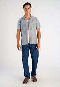 Koszula w paski z krótkim rękawem w kolorze jasnoniebieskim i białym, zapięta na guziki, zestawiona z ciemnoniebieskimi spodniami dżinsowymi i brązowymi butami. Czysty, casualowy wygląd.
