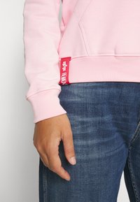 Felpa rosa con polsini a costine, dotata di un'etichetta rossa sul lato. Indossata con jeans in denim blu, che mostrano un taglio casual e rilassato.