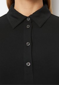 Detailní záběr černé košile s límečkem, se čtyřmi černými knoflíky zapnutými podél předního zapínání, kterou nosí osoba.