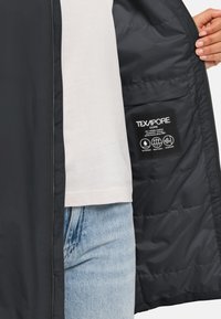 Veste imperméable noire avec doublure isolante, présentant une texture lisse, une fermeture à glissière et une étiquette indiquant la technologie TEXAPORE.
