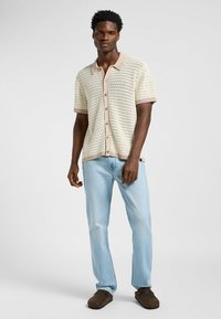 Kortärmad, strukturerad stickad skjorta i krämfärg med beige detaljer, knappstängning, ihop med ljusblå denimjeans och mörkbruna skor.