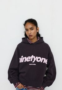 Tummanharmaa ylisuuri huppari, jossa vaaleanpunainen teksti "ninetyone" ja "visual studies". Alla valkoinen ja burgundinsävyinen raidallinen kerros, resorit hihansuissa.