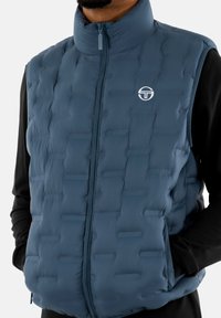 Gilet imbottito blu con colletto alto, chiusura a zip full, e logo grafico. Materiale testurizzato con motivi orizzontali e verticali. Maniche lunghe nere sotto.