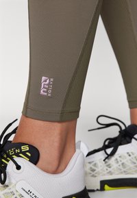 Leggings atletici a coste color verde oliva con un piccolo logo rosa. Indossati con scarpe da ginnastica bianche e nere caratterizzate da un motivo testurizzato e accenti vivaci.