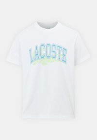 Balta kokvilnas T-kreklu ar zilu un zaļu "LACOSTE" grafisko tekstu priekšpusē un krokodila akcentu zem tā. Standarta apaļā kakla dizains.