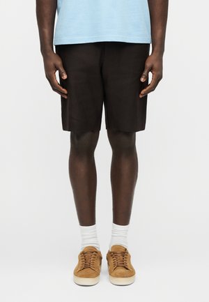 Persoon in zwarte shorts, witte sokken en bruine suède sneakers, staand tegen een effen witte achtergrond.