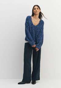 Niebieski, puszysty sweter z dekoltem w serek, o luźnym kroju i teksturowanej powierzchni. W zestawie z szerokimi, ciemnymi spodniami oraz czarnymi butami.