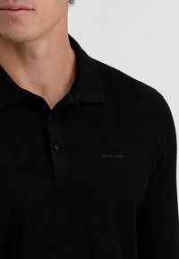 Zwarte polo gemaakt van textuurstof met een visgraatpatroon, voorzien van een drieknoopsluiting en een subtiele logo op de borst.