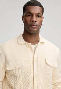 Vêtement d'extérieur tricoté couleur crème avec un col, présentant une texture en maille torsadée au centre et deux poches poitrine boutonnées.