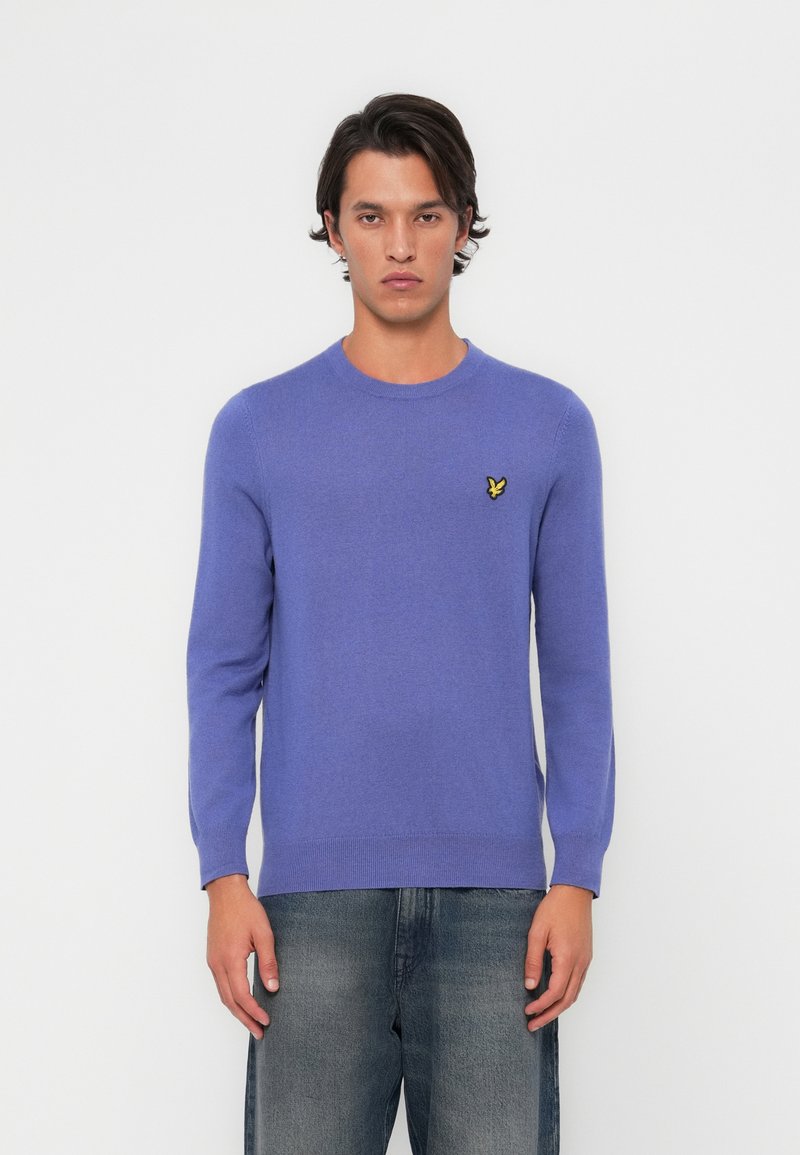Lila Pullover mit langen Ärmeln, rundem Ausschnitt und einem kleinen gelben Schmetterlingslogo auf der Brust. Weicher Stoff und figurnahes Design.