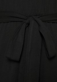 Ceinture en tissu noir nouée de manière soignée avec des extrémités lâches sur un fond de vêtement noir texturé.