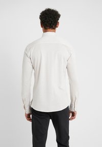 Camicia beige chiaro a maniche lunghe con colletto a bottoni, texture liscia e orlo curvo. Presenta polsini con bottone e design semplice.