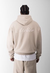 Sudadera beige con capucha y la palabra "Atelier" bordada en la parte posterior. Tiene un corte holgado, puños acanalados y una camiseta blanca de capas visible debajo.