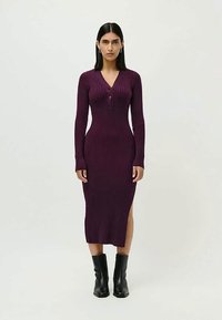 Nais, kes kannab sobivat, ribaga pikkade varrukatega lillat kleiti, millel on nupud rinna piirkonnas, külgauk ja mustad pahkluudega saapad, seismas ühtlase tausta ees.