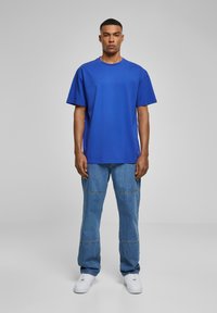 Blå bomulls T-skjorte med korte ermer og rund hals, kombinert med lyseblå jeans med sømmer. Modellen har på seg hvite joggesko.