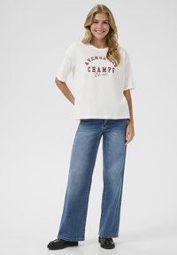 T-shirt crema oversize con la scritta "AVENUE" e "CHAMPS" in testo marrone, abbinato a jeans di denim blu a gamba larga e scarpe nere chunky.
