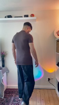 Brown fitted t-shirt with short sleeves, paired with loose dark pants. Background features colorful wall lights and a textured area rug.