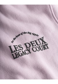 Ροζ φούτερ με μαύρο κείμενο: "Για την αγάπη της Μεγάλης Μήλου" και "LES DEUX LEGACY COURT." Μαλακό, υφασμάτινο υλικό.