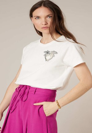 Femme aux longs cheveux bruns portant un t-shirt blanc avec un motif de palmier et un pantalon taille haute magenta avec une ceinture nouée, main dans la poche.