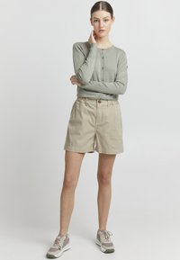 Lys grønn, langermet cardigan med knapplukking, kombinert med beige shorts med frontknapp og lommer, fullført med grå sneakers.
